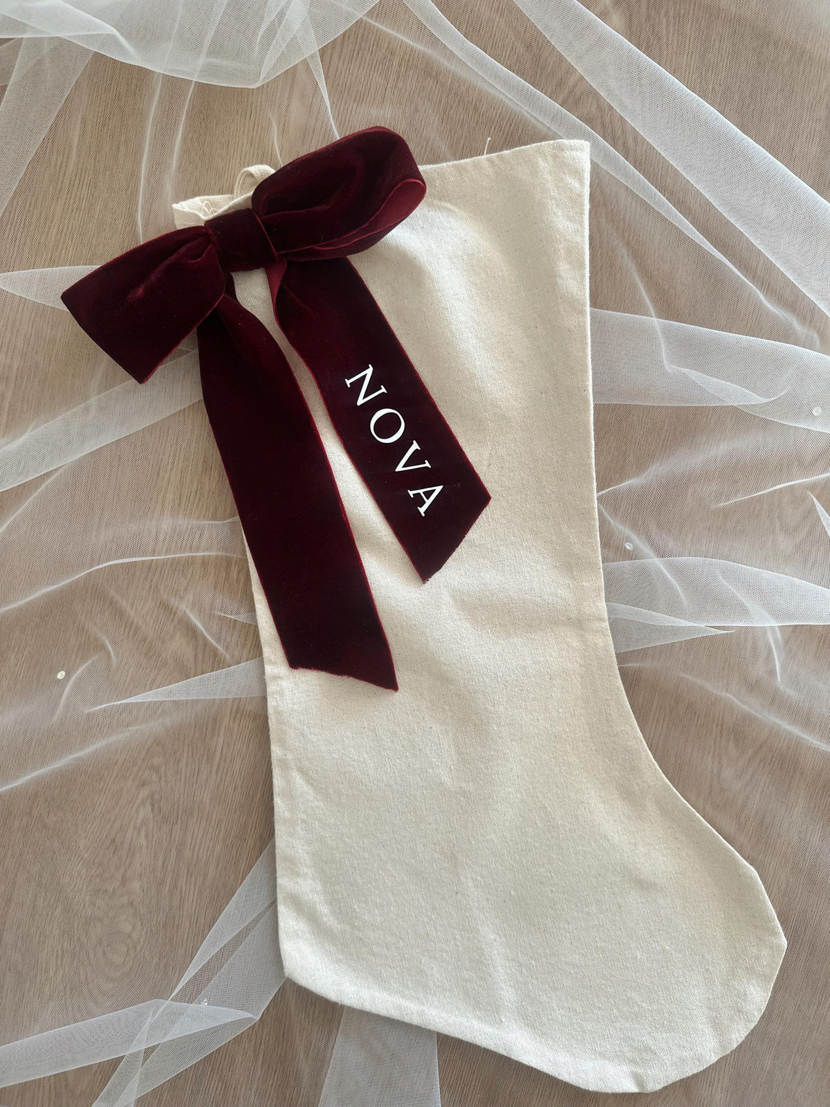 White Linen stocking with a burgundy velvet ribbon and 'NOVA' text on a sheer fabric background