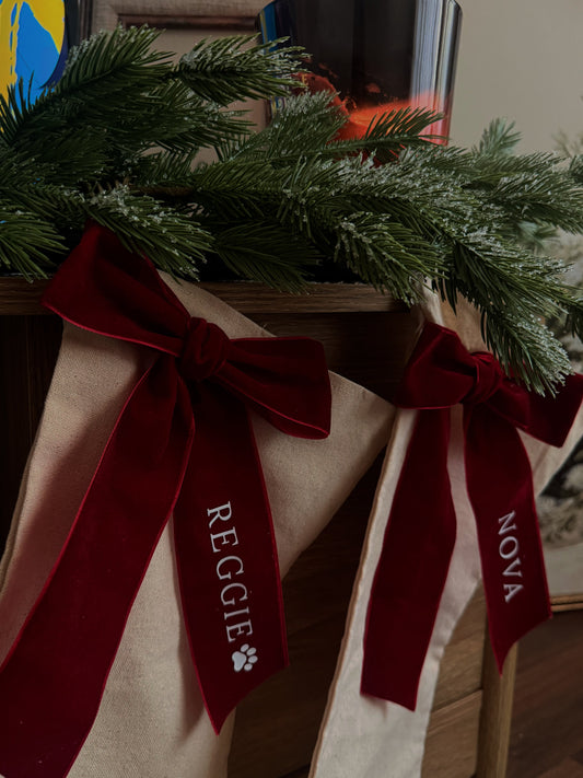 Two cream-colored Christmas stockings with large dark red velvet bows and personalized names 'REGGIE' and 'NOVA' printed in white, hanging on a wooden table decorated with green pine branches.