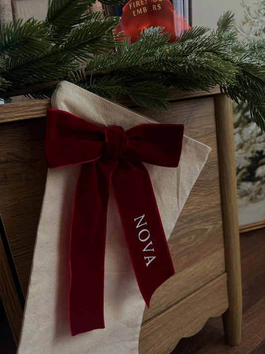 A cream-colored Christmas stocking with a large dark red velvet bow and the name 'NOVA' embroidered in white, hanging on a wooden cabinet next to green pine branches.